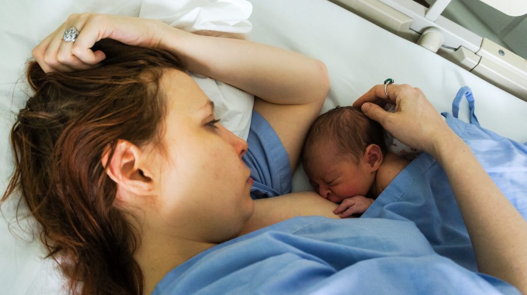 A mother lying in a hospital bed breastfeeds her newborn for the first time