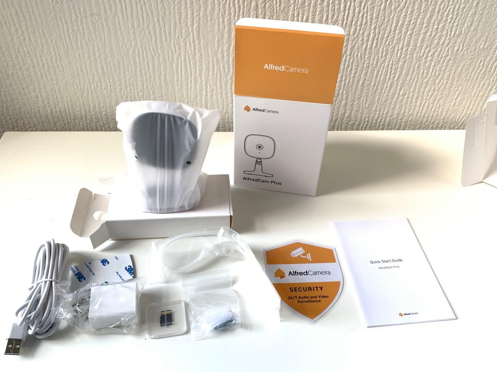 The packaging box and contents of AlfredCam Plus, which include the camera, USB C cable, power adapter, mounting sticker, alignment sticker, plastic cable ties, screws and wall plugs, as well as a 64 GB microSD card.